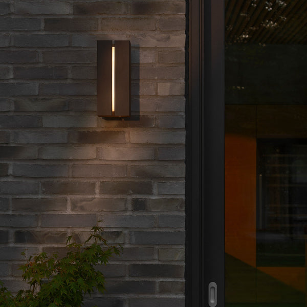 Black outdoor wall light with a vertical light strip on a dark brick wall next to a glass door