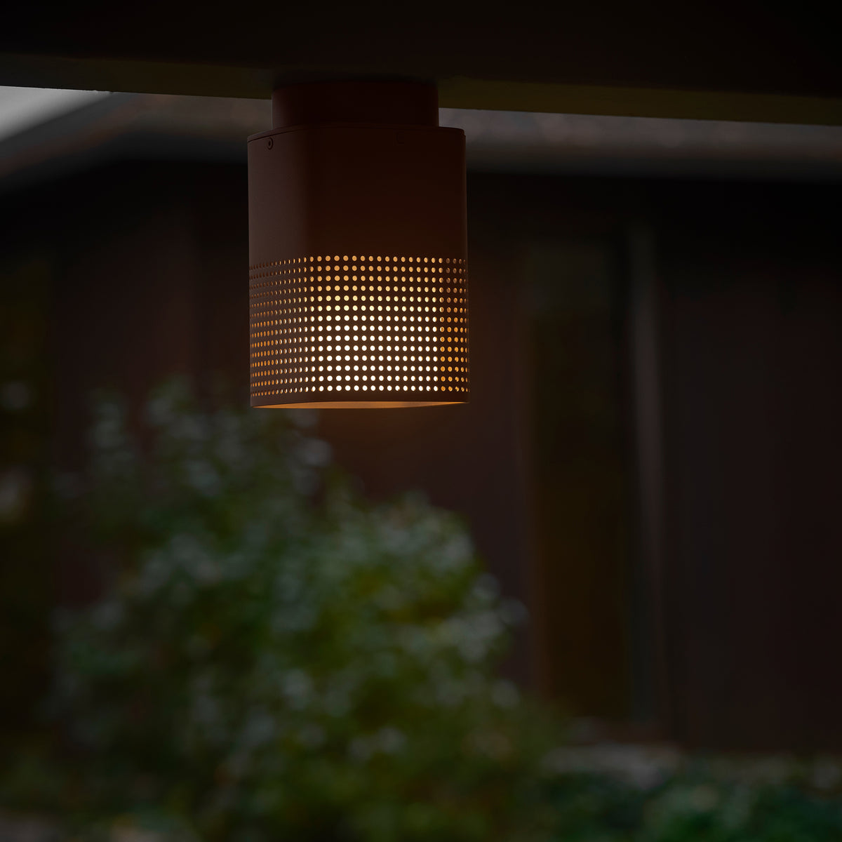 Perforated cylindrical outdoor light fixture with warm glow, mounted under a dark overhang, with blurred foliage below