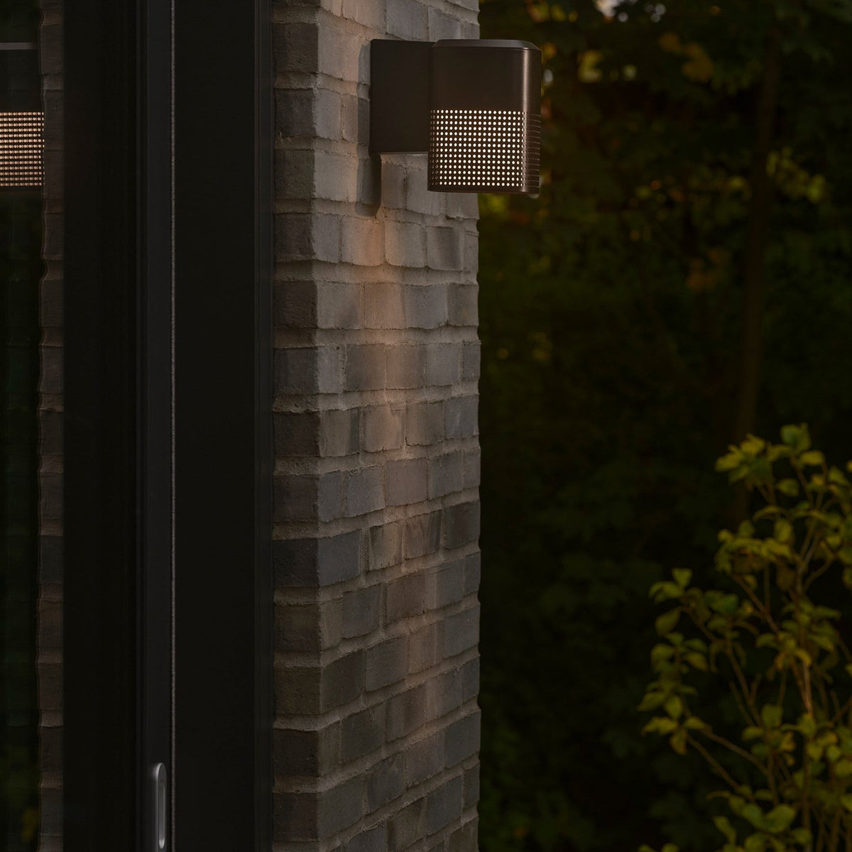 Black outdoor wall light with perforated front, casting light on a brick wall at dusk