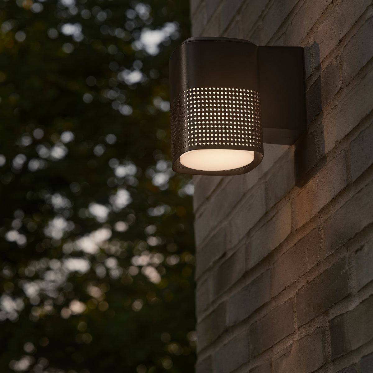 Black outdoor wall light with perforated shade on a brick wall, green foliage in the background