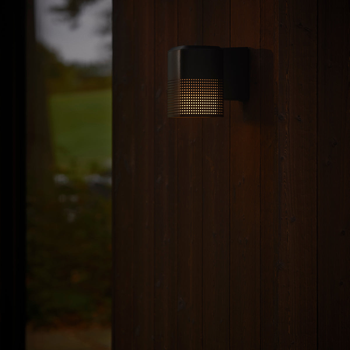 Black outdoor wall light with a perforated design, casting a warm glow on a dark wooden wall