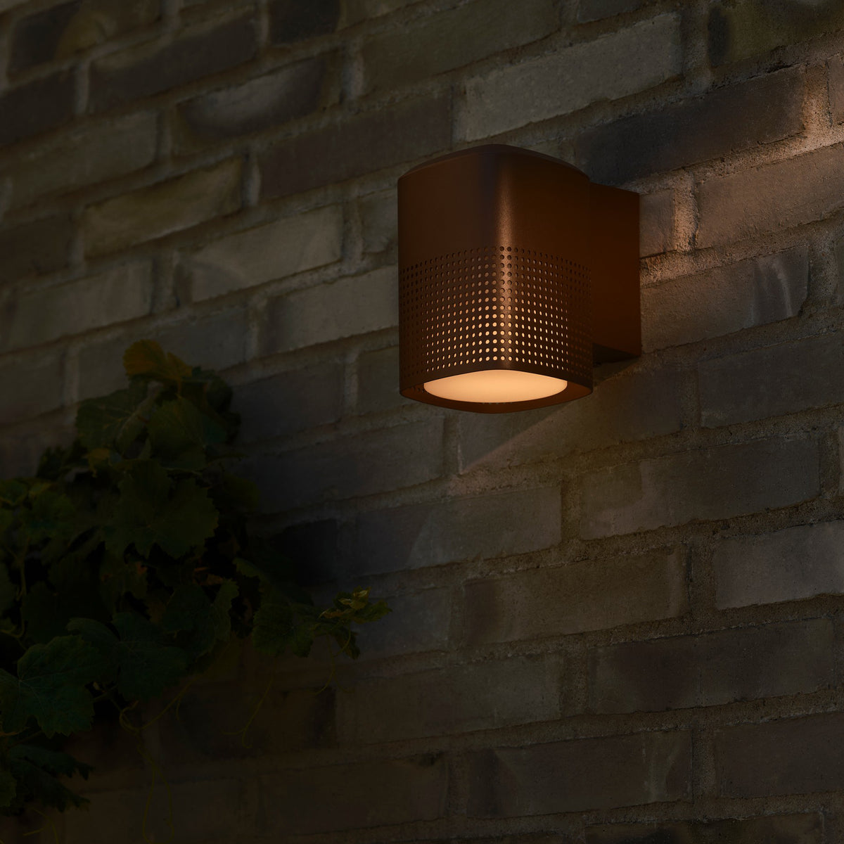 Brown outdoor wall light with perforated sides and a warm glow, mounted on a brick wall next to green foliage