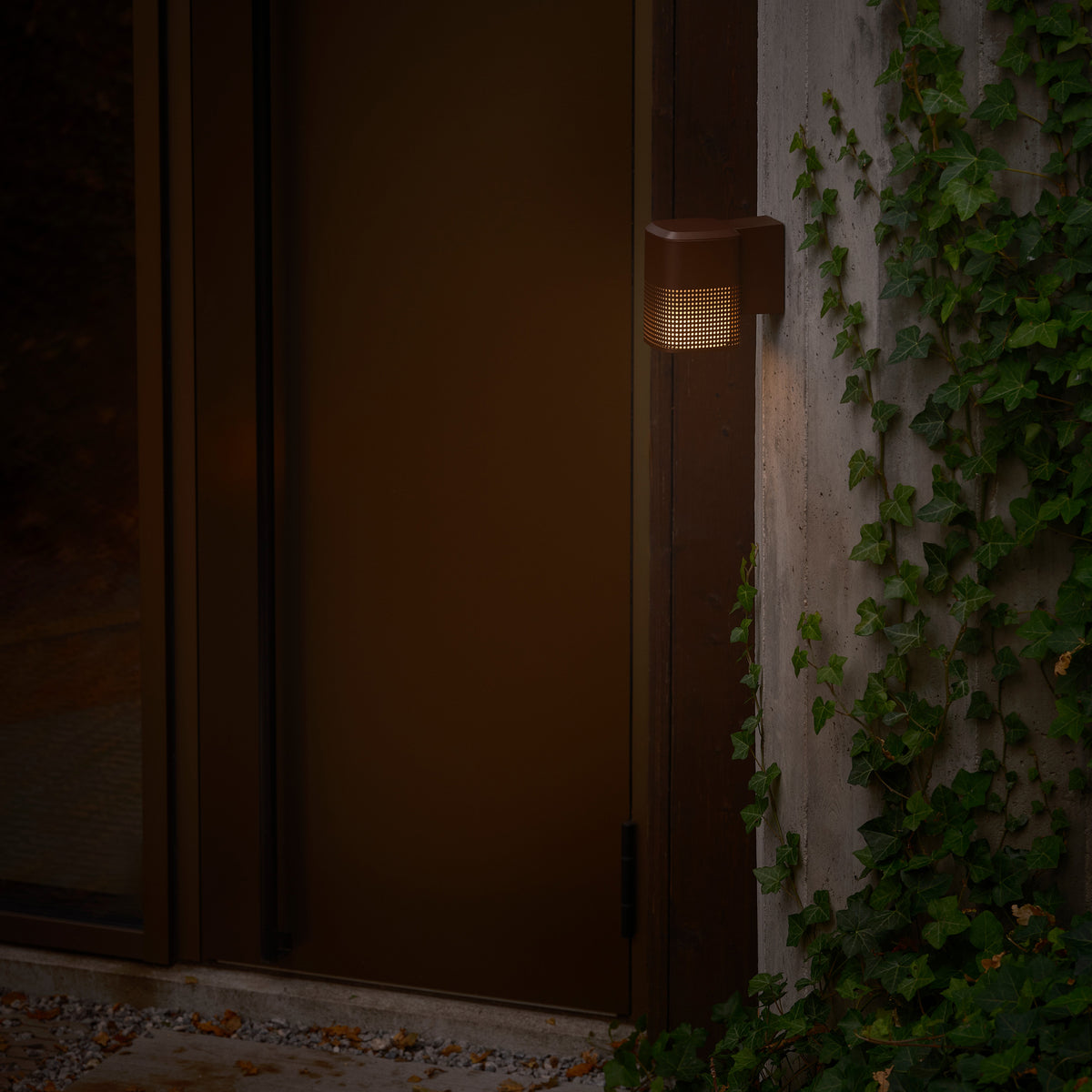 Brown outdoor wall light with a perforated shade, illuminating a concrete wall with ivy and a dark door