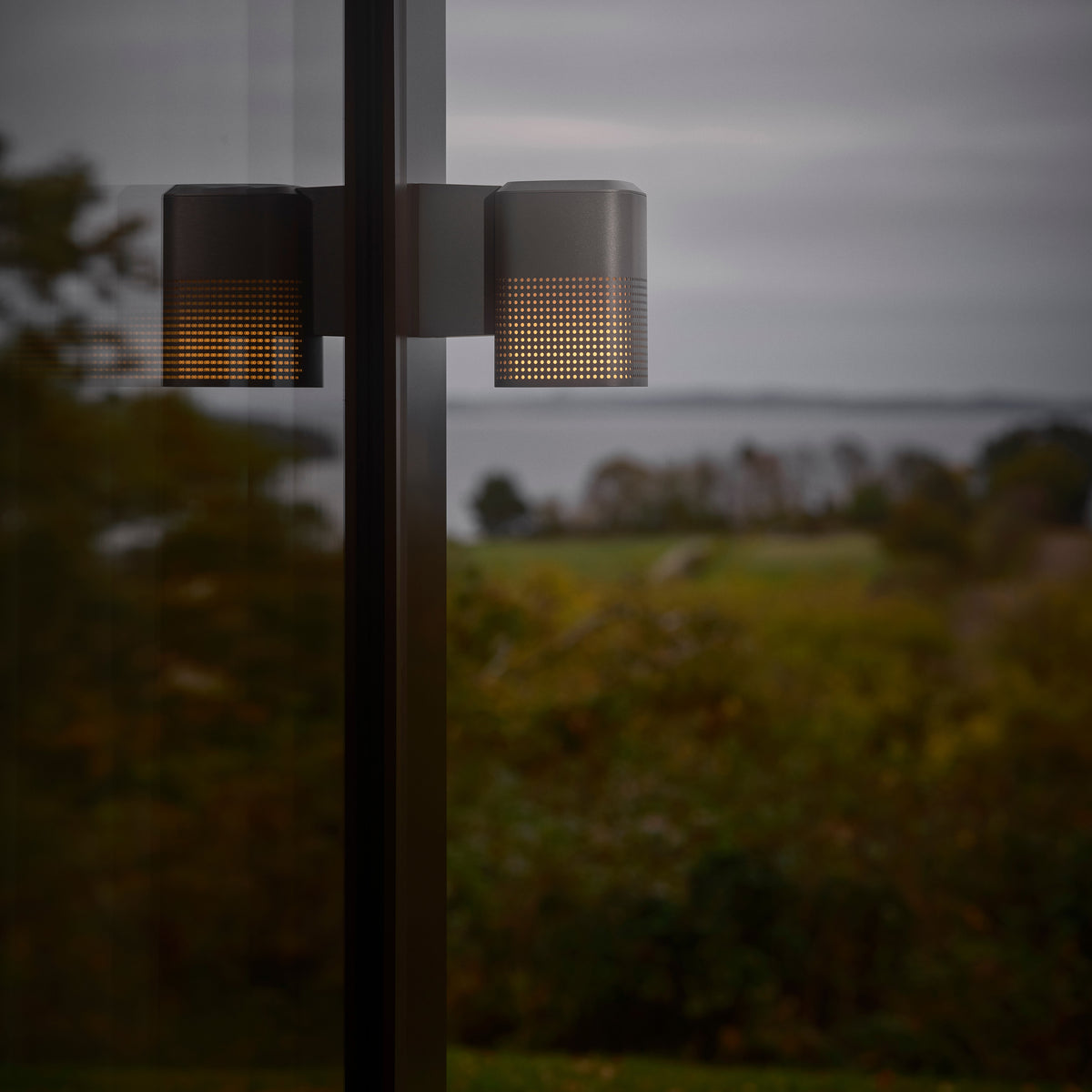 Two grey outdoor lights with perforated shades, illuminating a dark landscape with a body of water