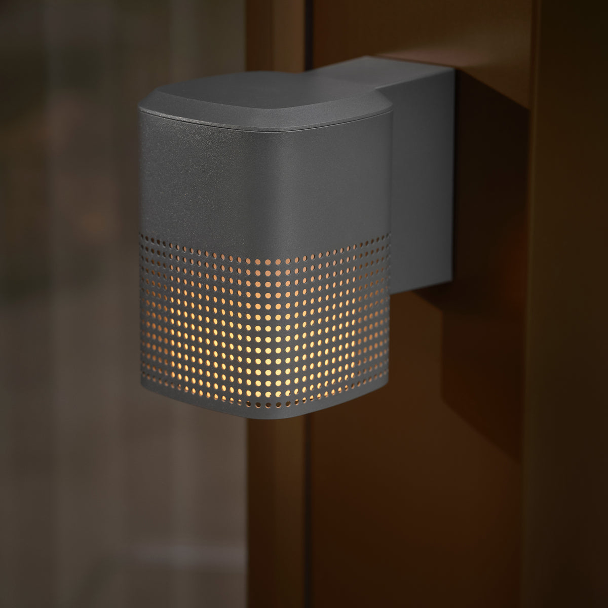 Dark grey outdoor wall light with a perforated pattern on the lower half, emitting a warm glow