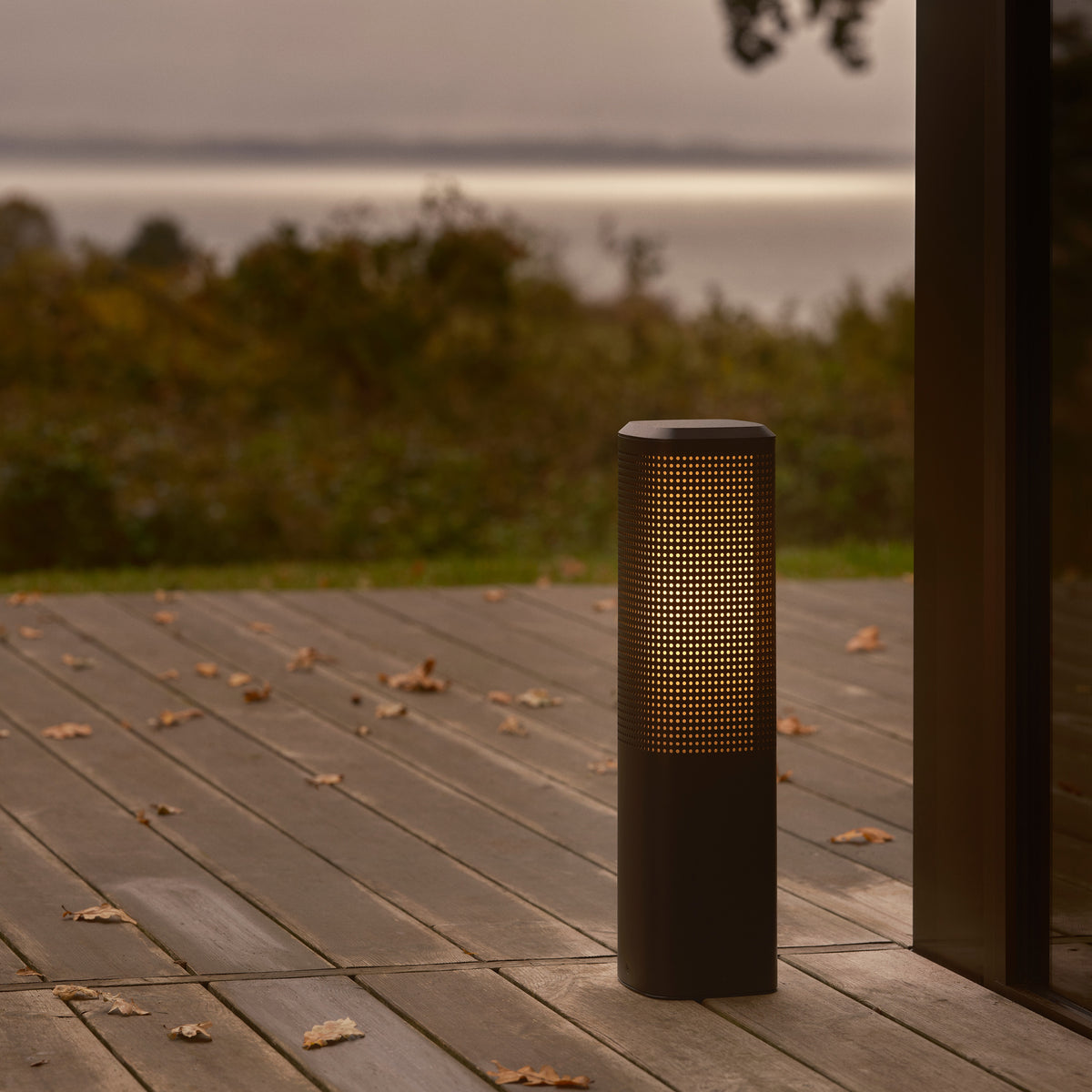 Dark grey cylindrical outdoor lamp with a perforated top half, emitting a warm glow, on a wooden deck