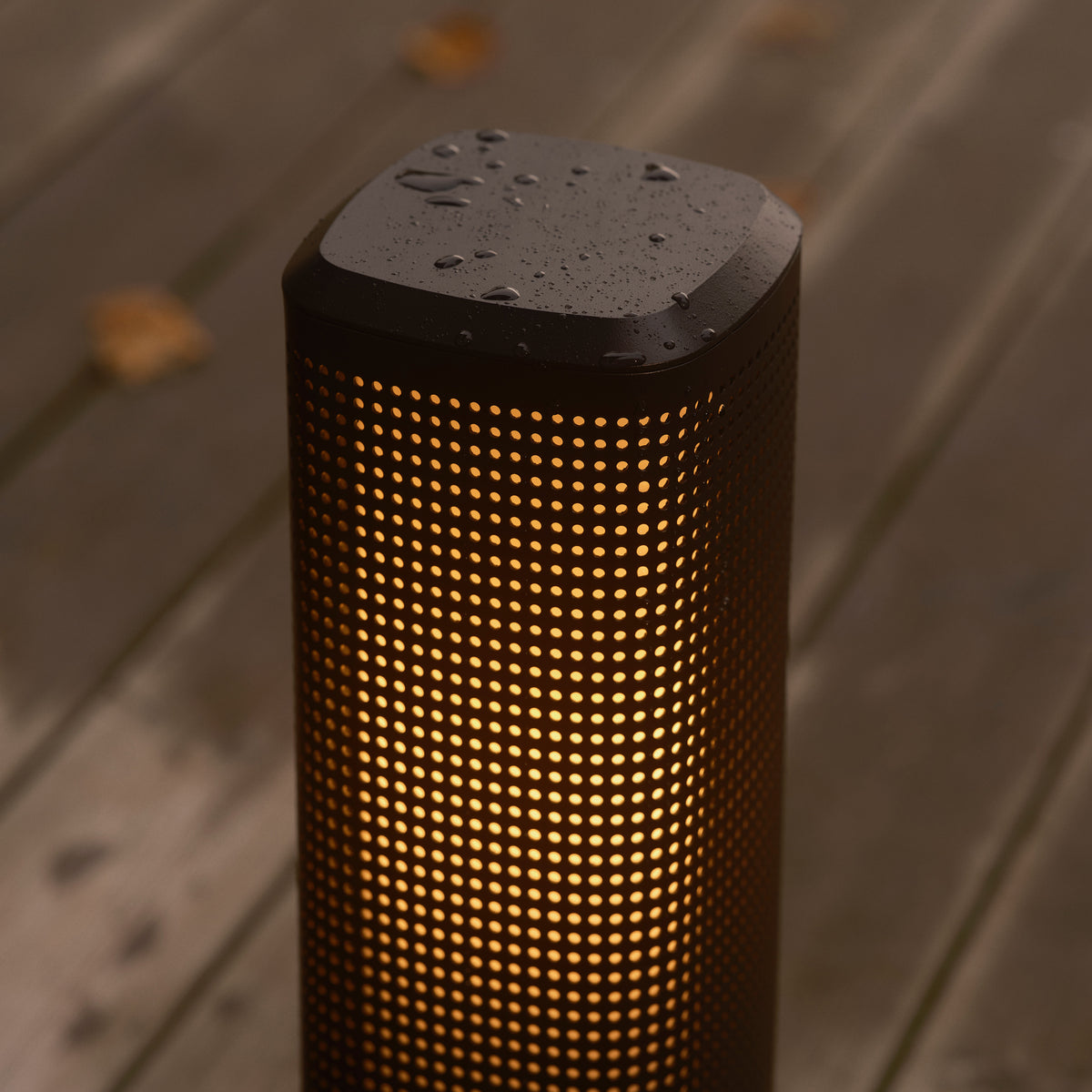 Black cylindrical outdoor light with small holes and warm glow, water droplets on top, on wooden deck