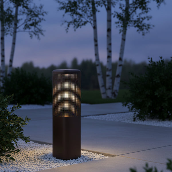 Brown cylindrical outdoor light with perforated top half, illuminating white pebbles and a concrete path at dusk