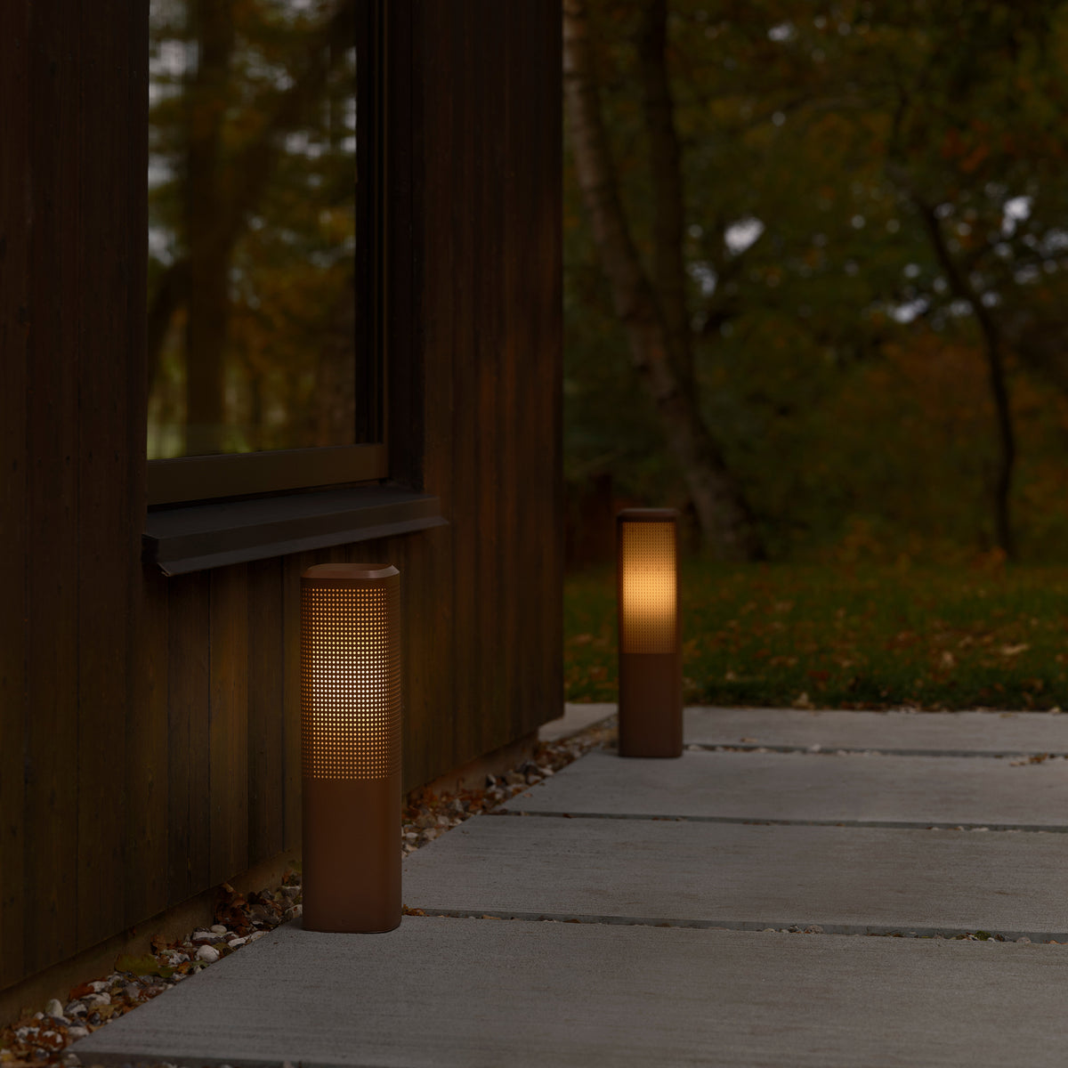 Two outdoor bollard lights illuminate a concrete path next to a wooden house with a window