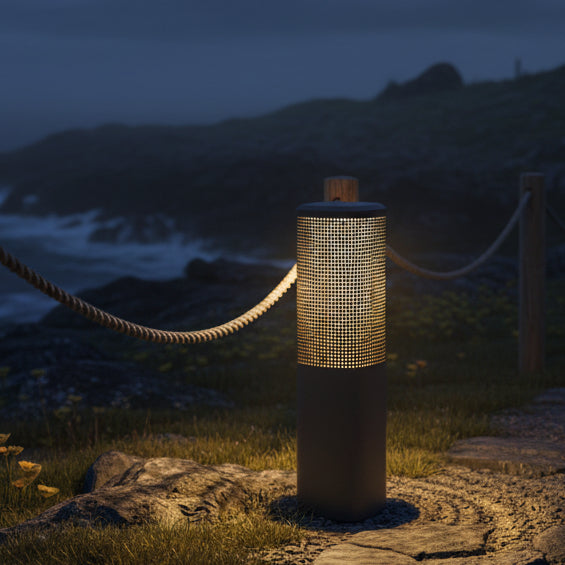 Bollard light with a perforated top section illuminating a path next to a rope fence and ocean