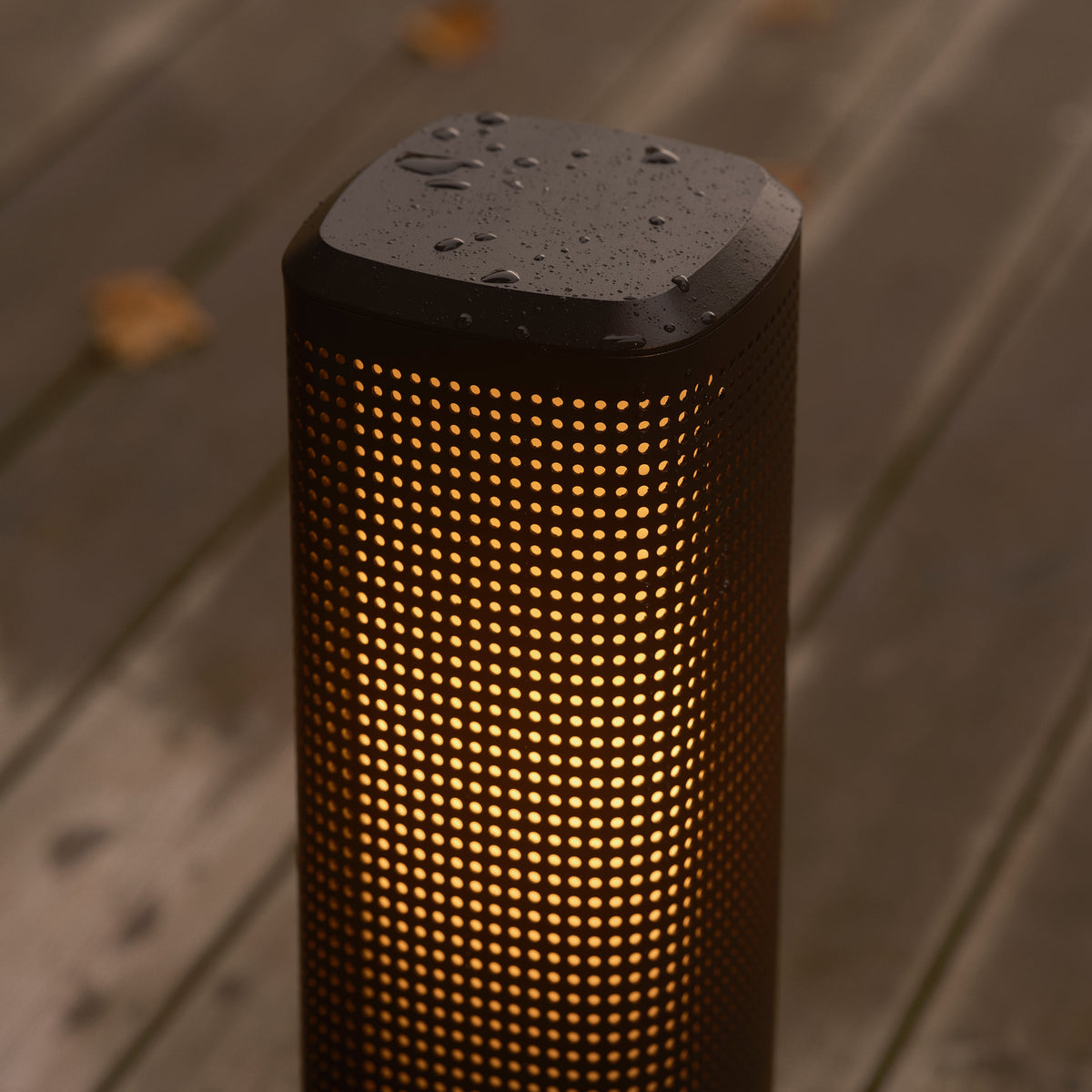 Black outdoor lantern with warm light and water droplets on top, on a wooden deck
