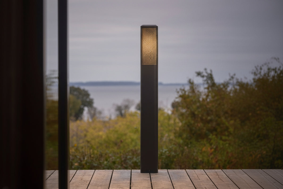 Tall dark grey outdoor light with a glowing patterned top, on a wooden deck overlooking a lake and trees