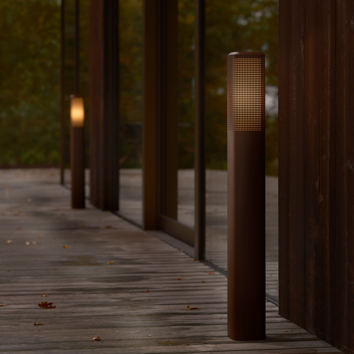 Two brown bollard lights with perforated tops illuminate a wooden deck next to a building with large windows
