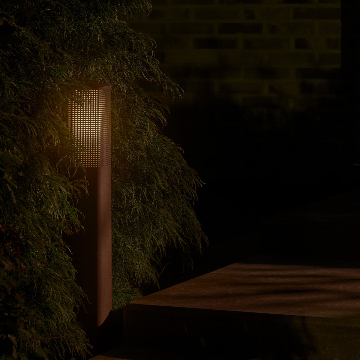 Outdoor bollard light with perforated shade illuminating a dark garden path next to green foliage