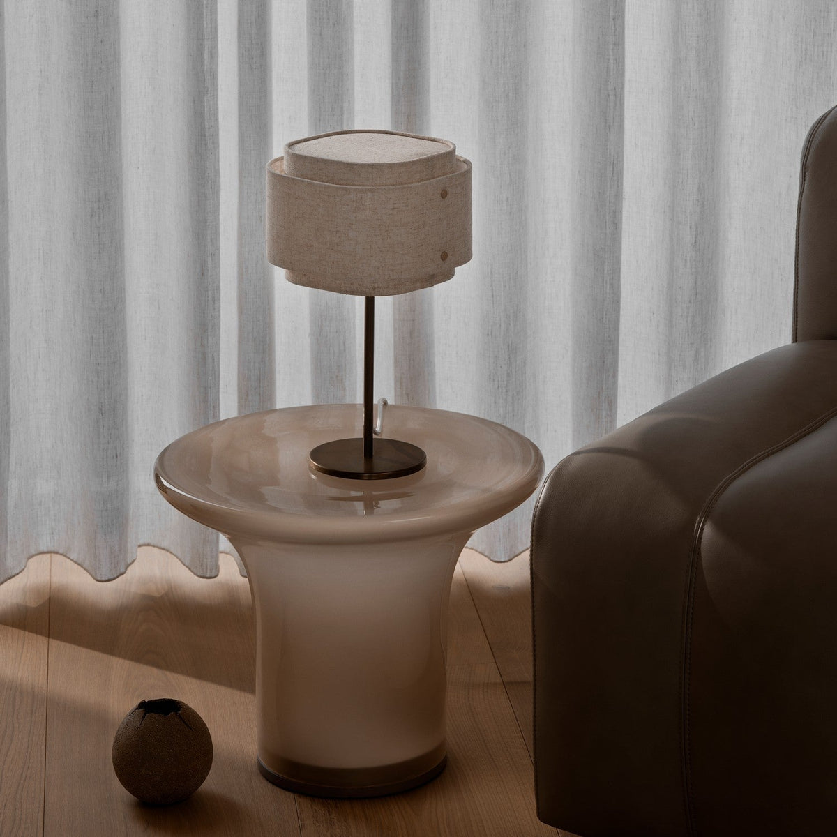 A table lamp with a beige fabric shade on a glass side table next to a brown leather sofa