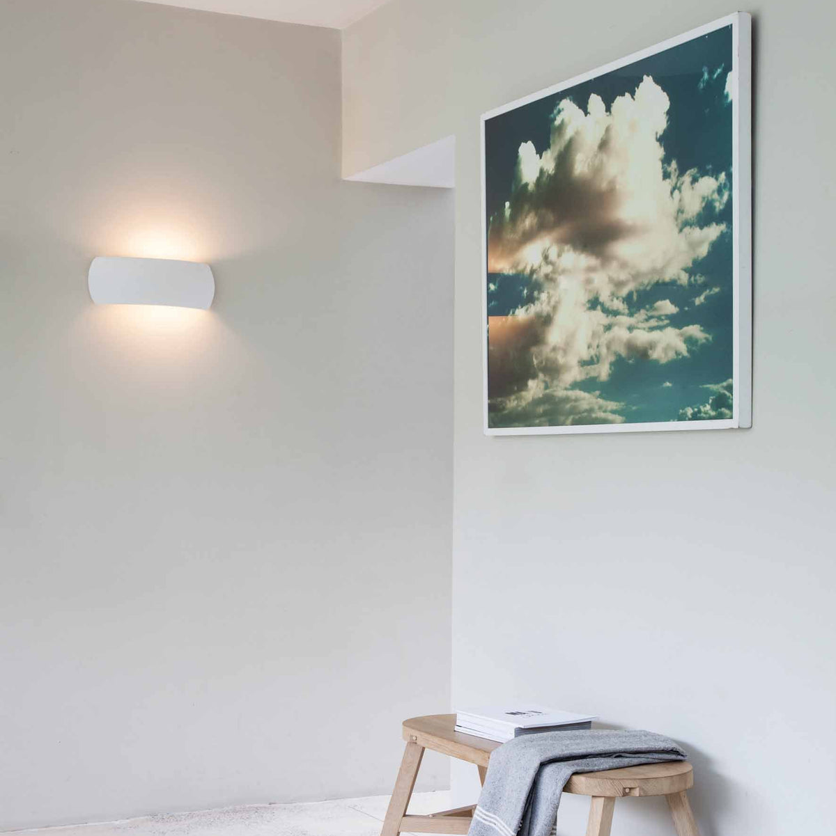 White wall light illuminating a light grey wall, with a wooden stool and a framed cloud print
