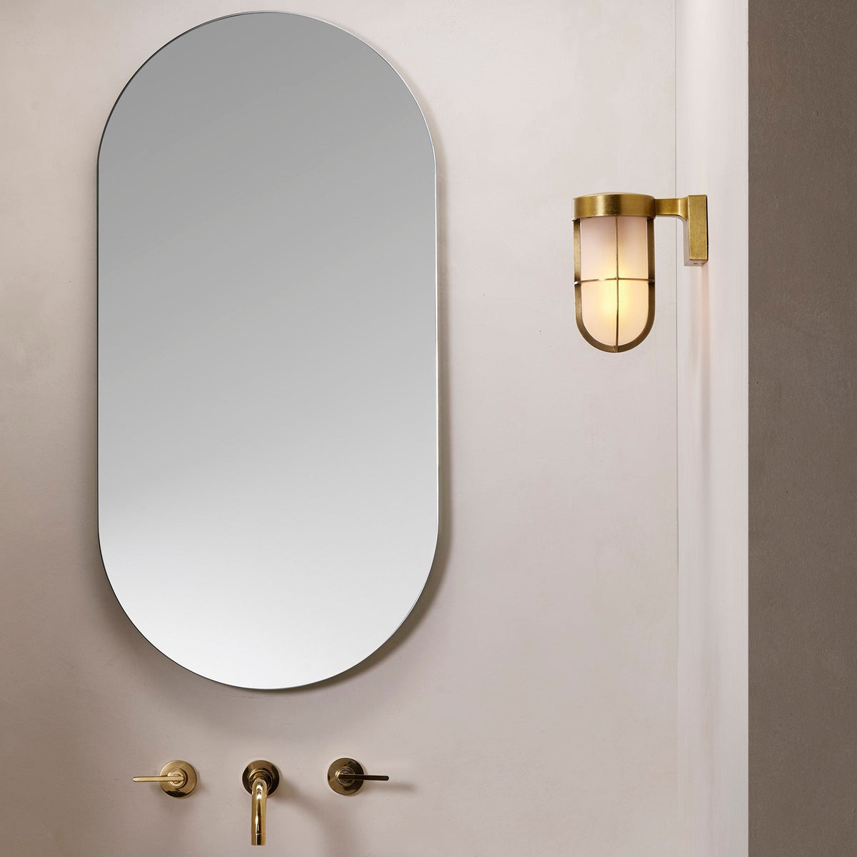 Oval mirror above three brass wall-mounted taps and a brass wall light