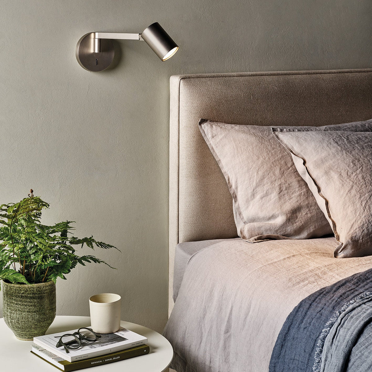 Bedroom with a bed, side table, plant, books, glasses, and a wall-mounted light