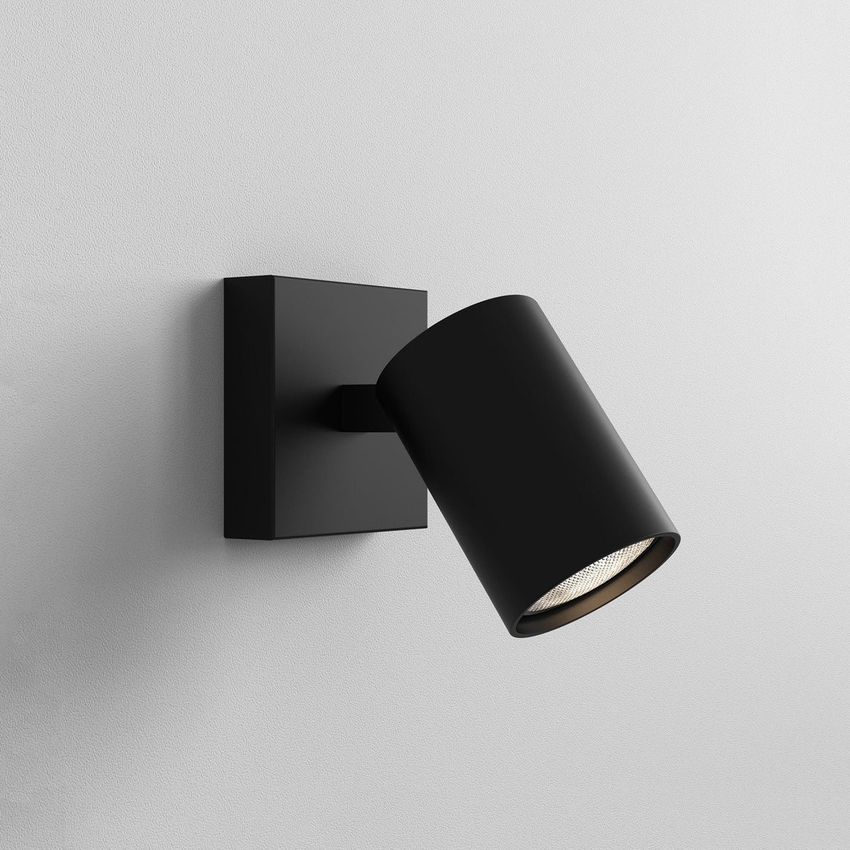 Black wall-mounted spotlight with a square base and cylindrical head, angled downwards, emitting a warm light