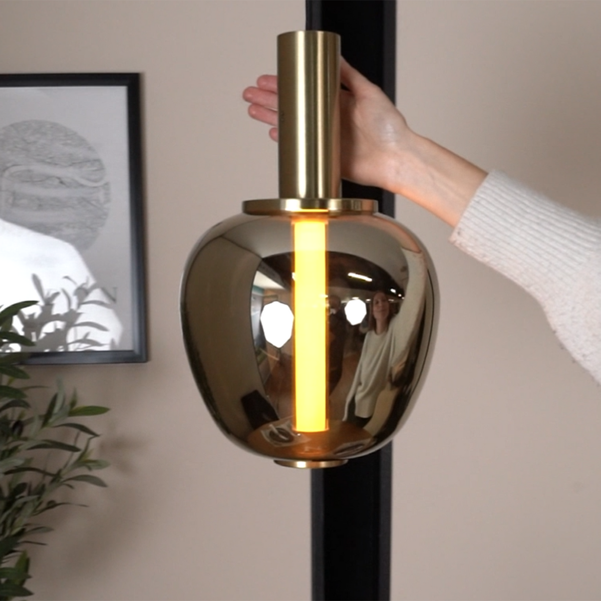 Hand holding a gold pendant light with a glowing yellow tube inside, reflecting a person and lights.