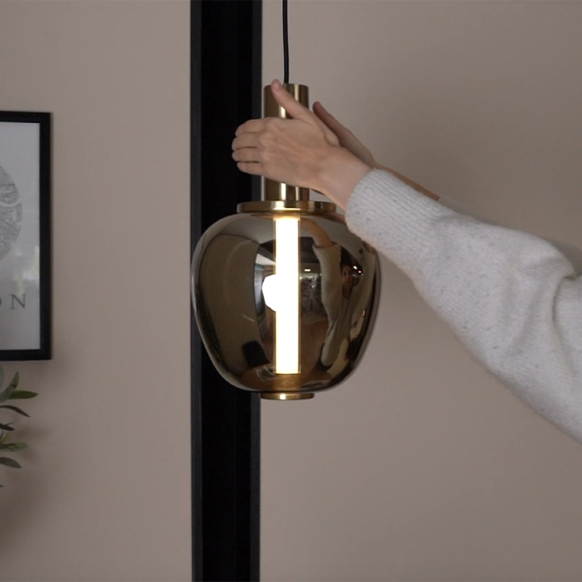 Hands adjusting a modern gold pendant light with a glowing vertical bulb.