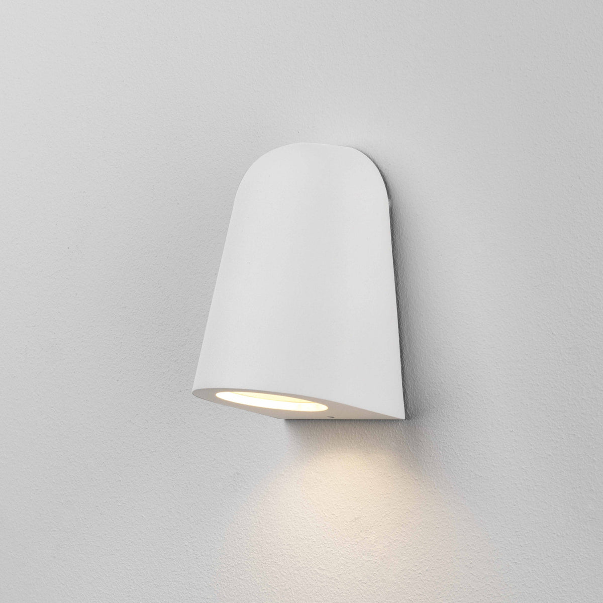 White wall light with a rounded top and an angled bottom, emitting warm light downwards onto a white wall