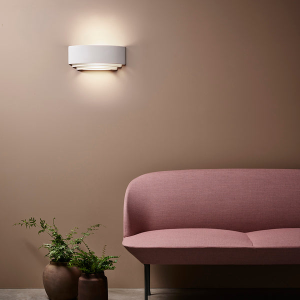 White wall light illuminating a brown wall above a pink sofa and two potted plants