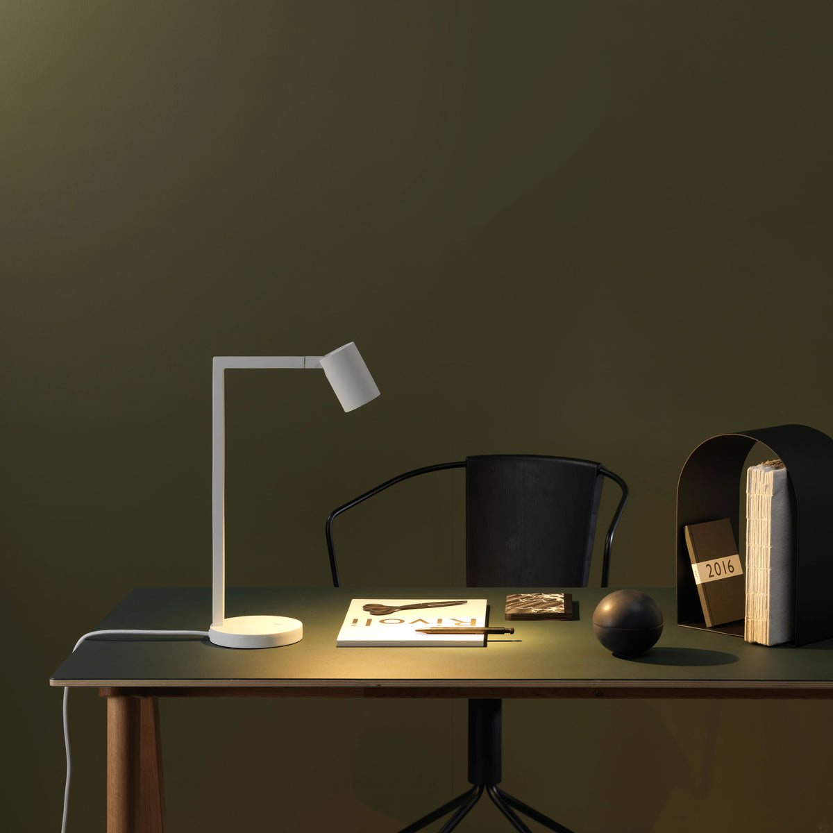 White desk lamp on a dark green desk with a black chair and books