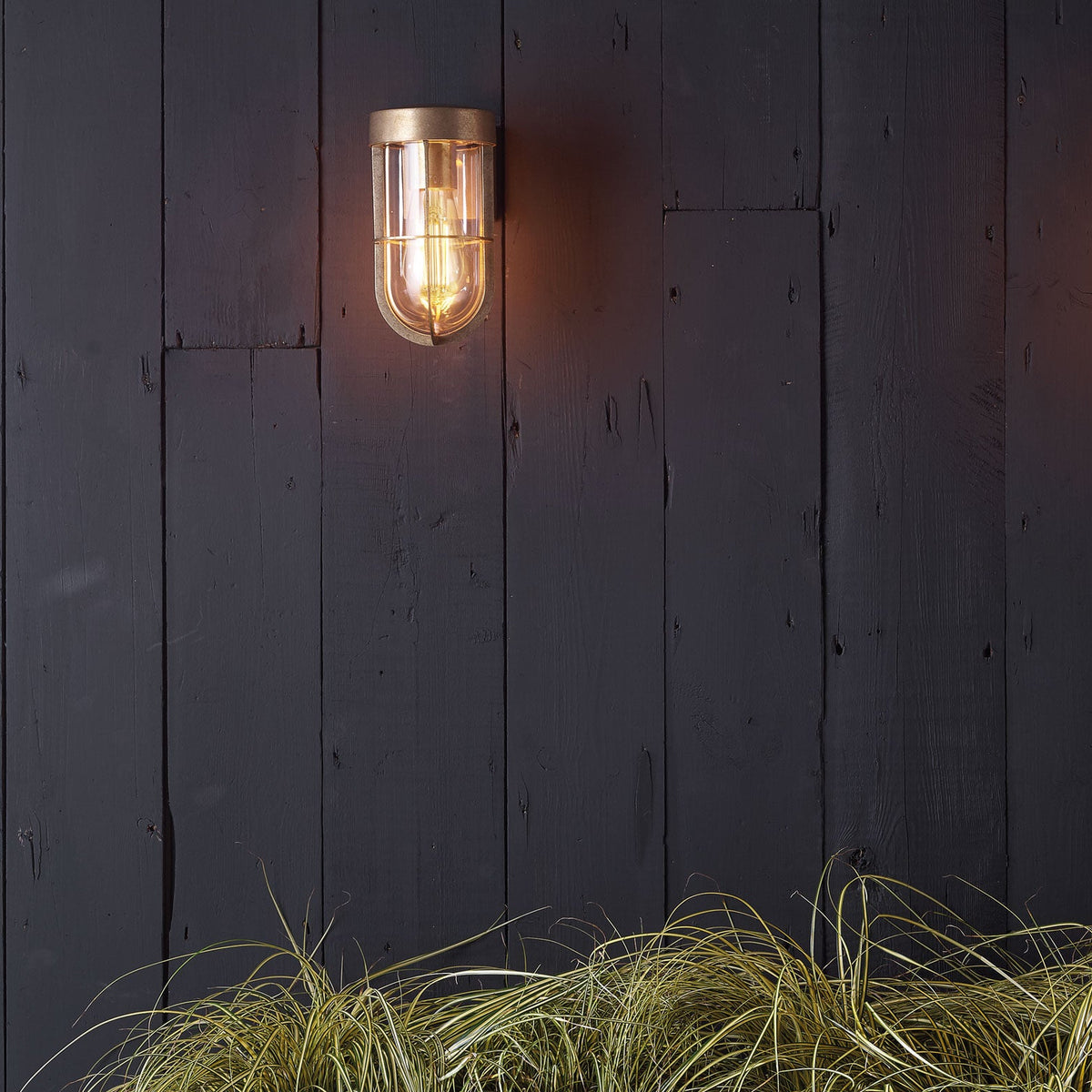 A brass outdoor wall light with a clear glass shade and exposed bulb, mounted on dark wooden planks