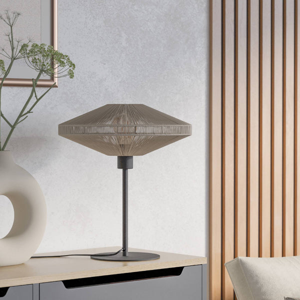 Table lamp with a woven shade and dark base on a wooden surface next to a white vase with greenery