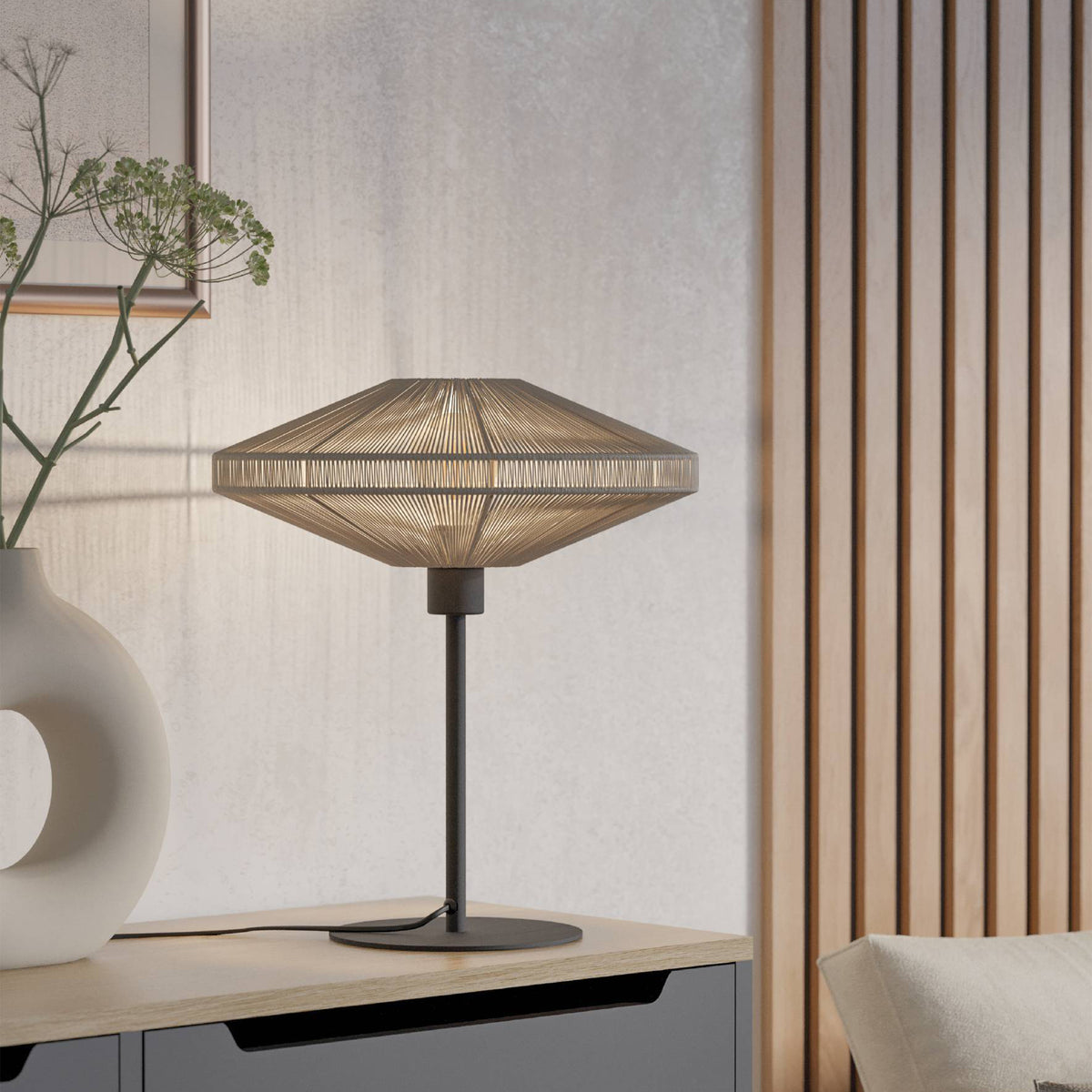 Table lamp with a woven shade on a wooden nightstand next to a vase with greenery