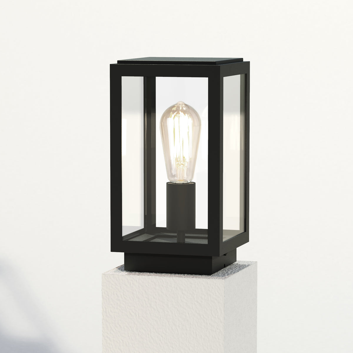 Black outdoor pillar light with clear glass panels and an exposed Edison bulb on a white textured base
