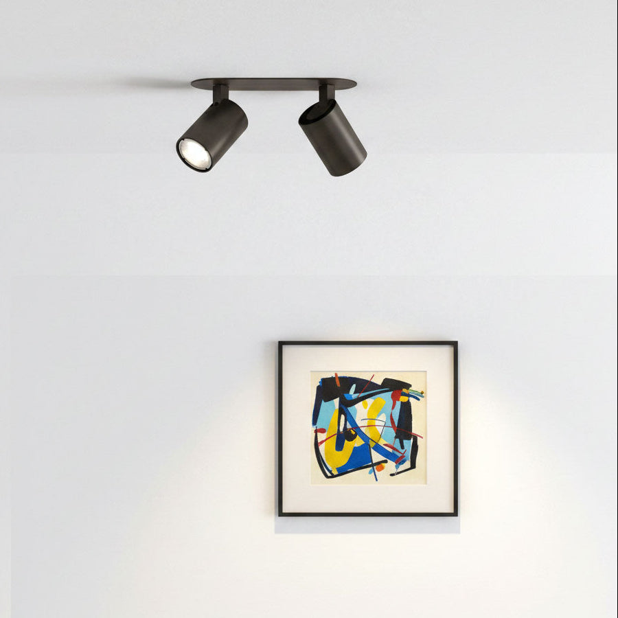 Two dark brown ceiling spotlights illuminating a framed abstract painting on a white wall