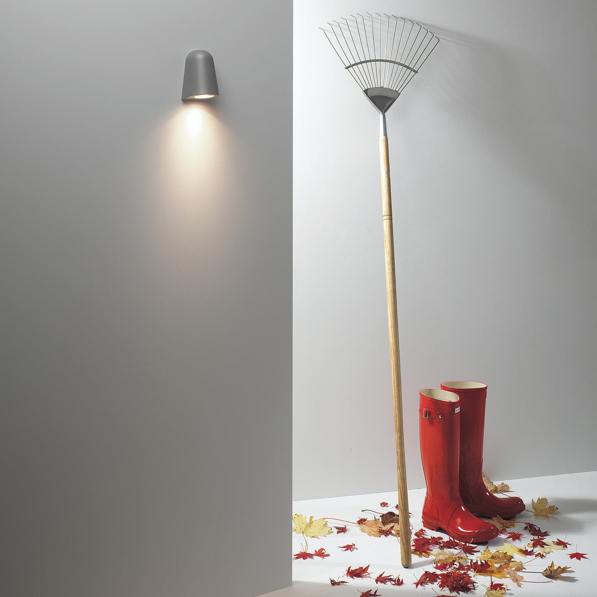 A grey wall light illuminates a corner with a rake, red boots, and autumn leaves