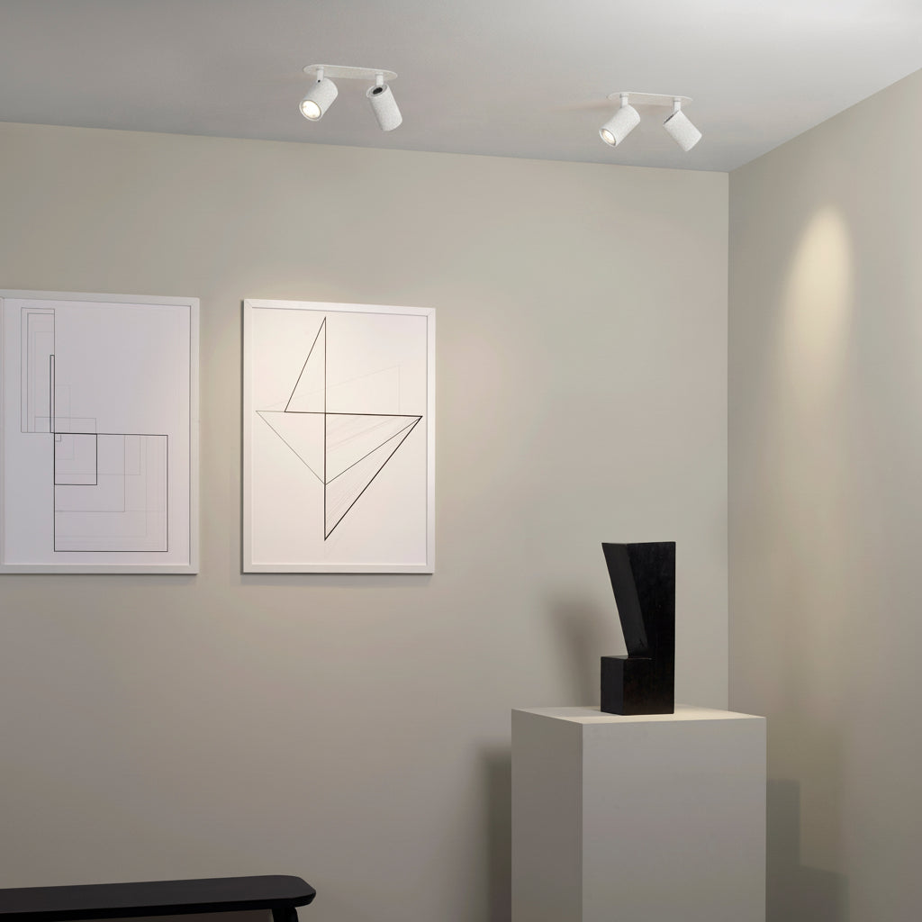 Two white ceiling spotlights illuminate a room with two framed geometric prints and a black sculpture