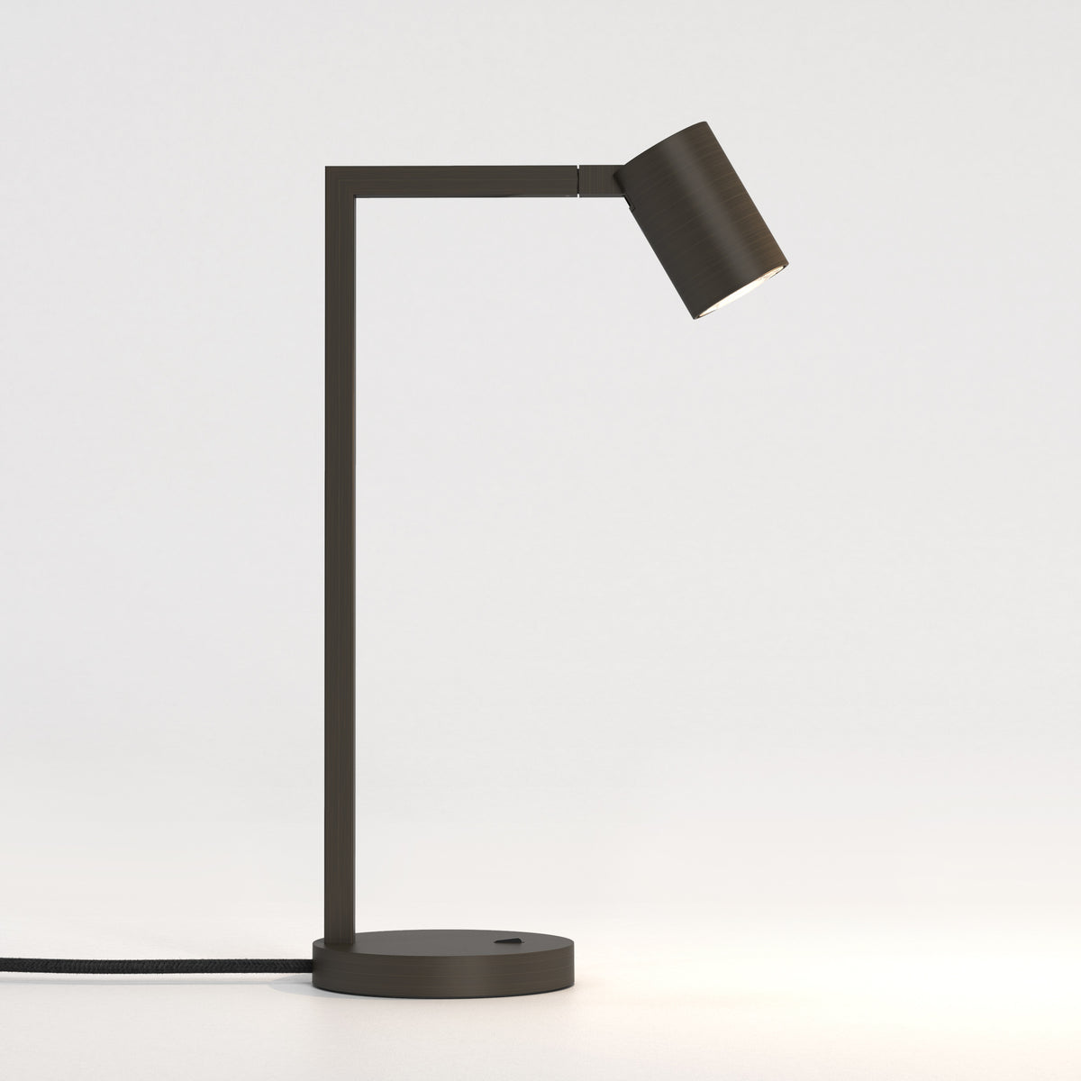 Dark bronze desk lamp with a cylindrical head angled downwards, emitting a warm glow onto a white surface