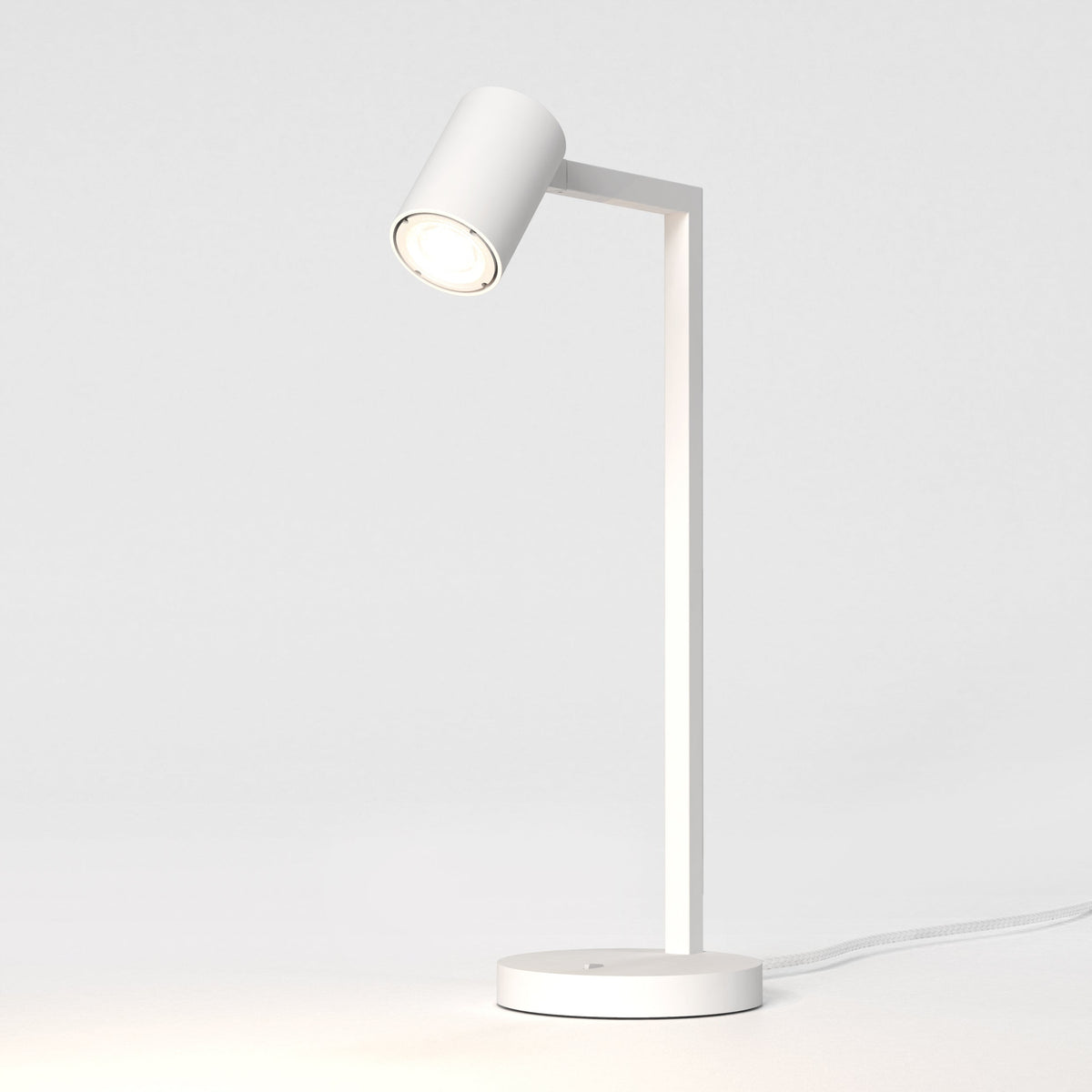 White desk lamp with a cylindrical head and a rectangular arm, illuminating a white surface