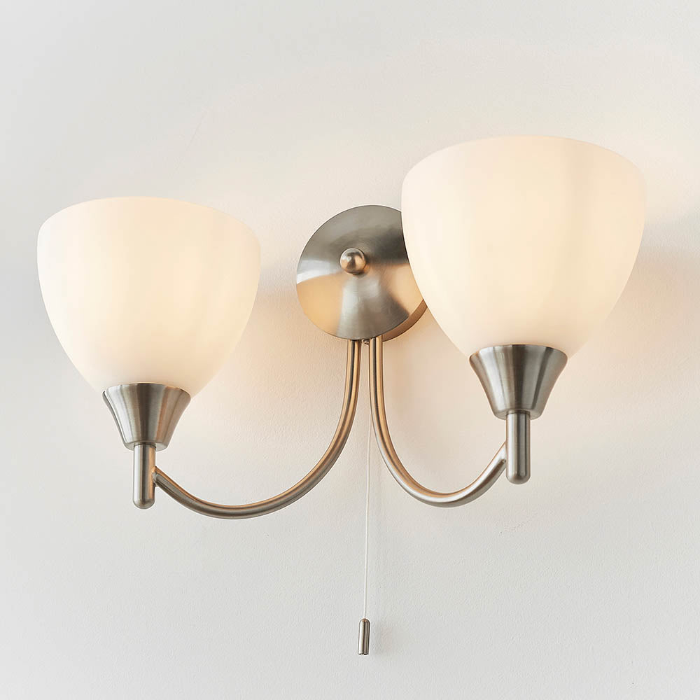 Two-light wall sconce with brushed nickel finish and frosted glass shades, illuminated on a white wall.
