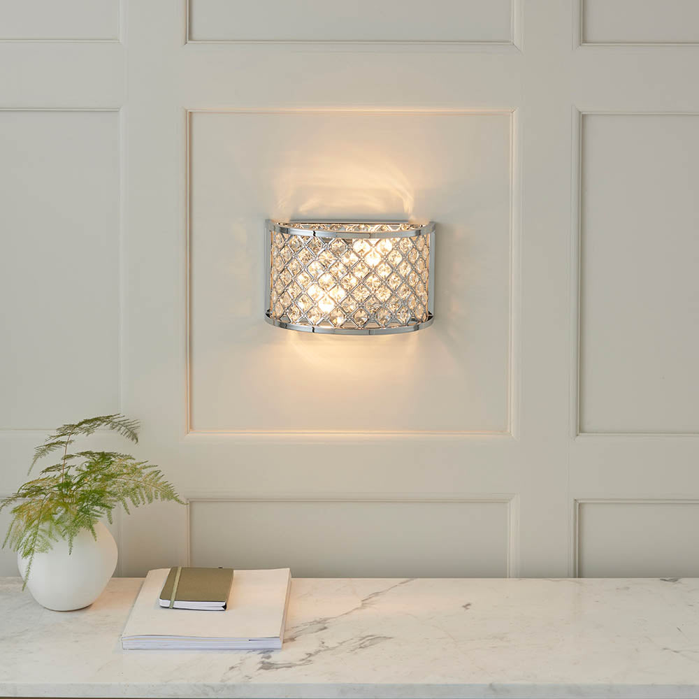 Chrome crystal wall light on a white paneled wall, illuminating a marble console table with decor.