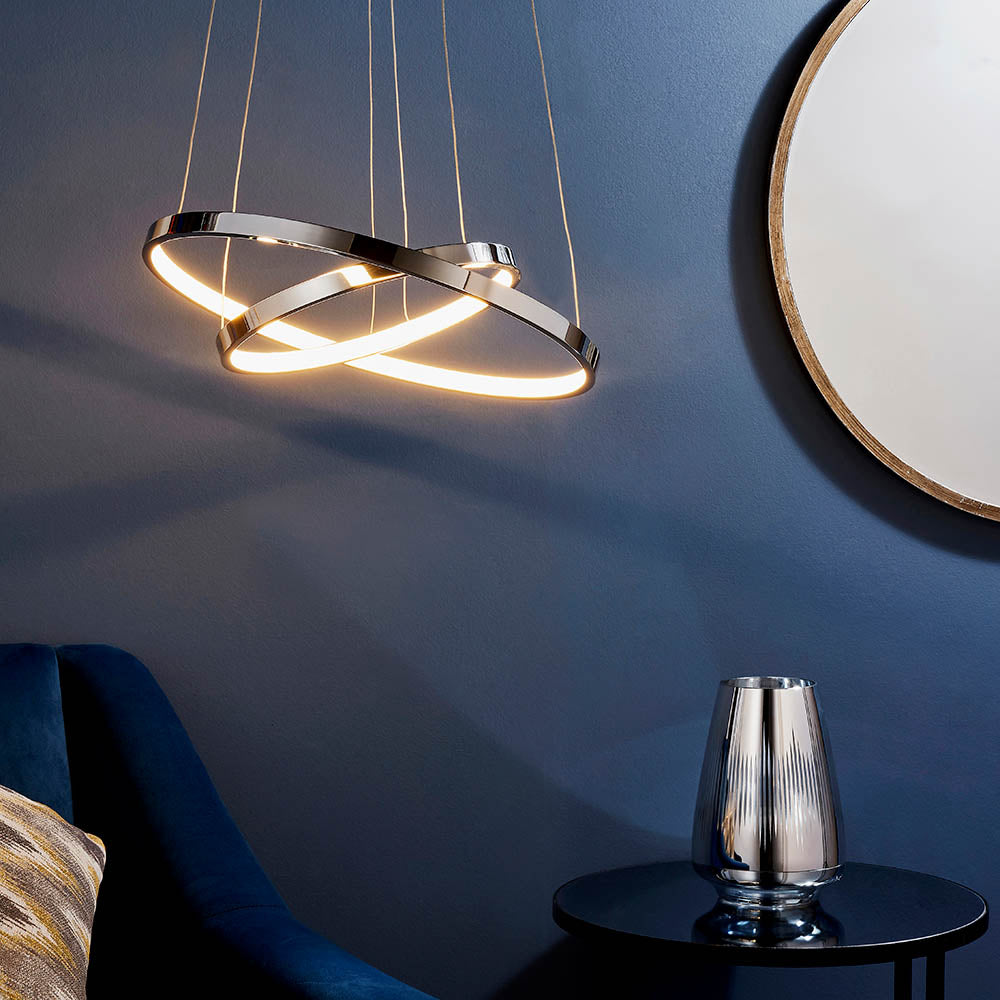 Chrome LED pendant light with two intertwined rings, illuminating a dark blue room with a gold-framed mirror.