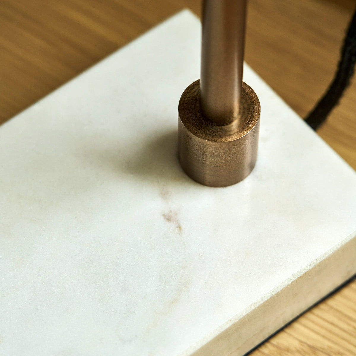 A close-up of a modern table lamp base featuring a white marble slab platform and a brushed bronze cylindrical metal stem, set on a light wood surface.