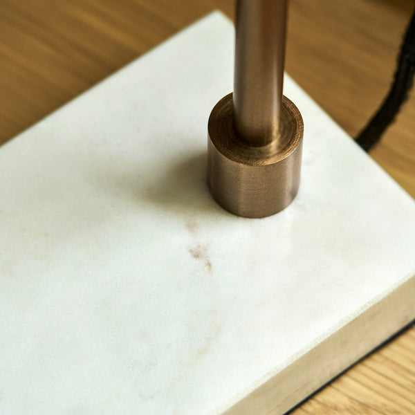 A close-up of a modern table lamp base featuring a white marble slab platform and a brushed bronze cylindrical metal stem, set on a light wood surface.