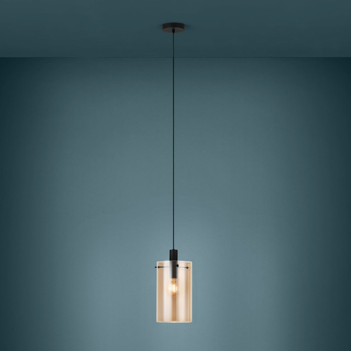 A modern pendant light with a cylindrical amber glass shade and a black fixture, hanging against a teal wall.