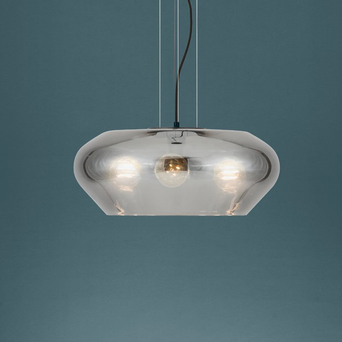 A modern, smoked glass pendant light with three visible bulbs, hanging against a dark teal background.