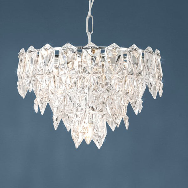 A modern chandelier with multiple clear, faceted crystal pieces hanging from a silver frame. It emits a soft glow.
