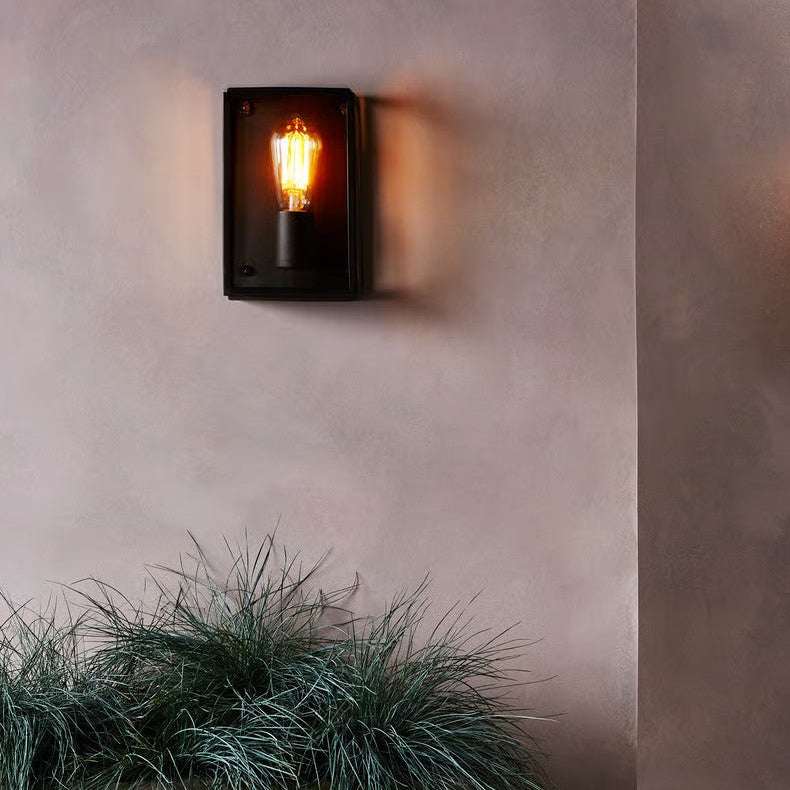 Black rectangular wall light with an exposed filament bulb, mounted on a textured wall above green plants