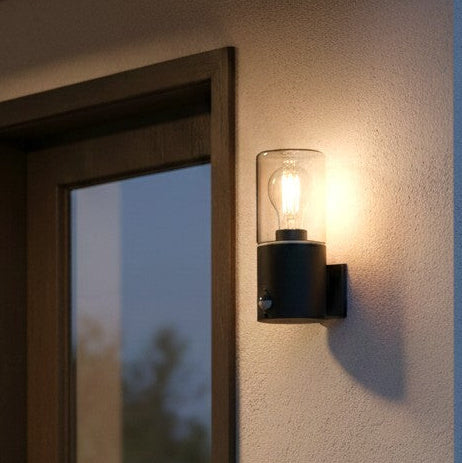 Black outdoor wall light with clear glass shade and visible bulb, mounted on a textured wall next to a door frame