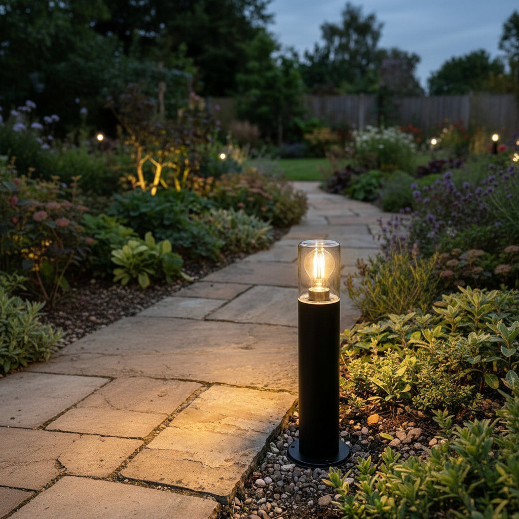 Black cylindrical outdoor light with a clear glass top and a glowing bulb, illuminating a garden path