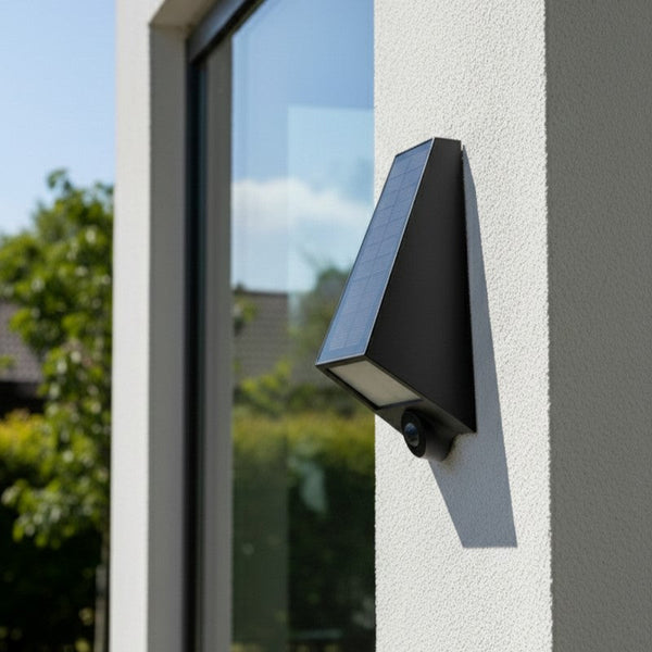 Black solar-powered security camera mounted on a white textured wall next to a large window