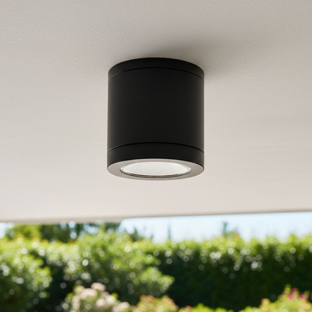 Black cylindrical outdoor ceiling light with a silver rim, mounted on a white ceiling with green foliage in the background